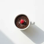 Chocolate brownie in a mug topped with a fresh raspberry and powdered sugar on a white background.