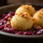 German Cabbage and Dumplings on dark ceramic plate with three fluffy potato dumplings topped with breadcrumb topping on bed of braised red cabbage