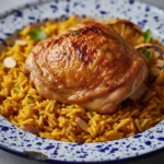 One-Pan Chicken and Rice Bake: Best Comfort Food That Your Family Will Love! 7 One-Pan Chicken and Rice Bake on blue speckled plate with golden-brown crispy chicken thigh on bed of yellow saffron rice with caramelized onions and fresh herbs
