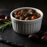Slow-cooked pork stew served in a white ramekin with tender pork chunks, carrots, herbs, and olives on a dark slate board.