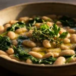 A rustic bowl of white beans and wilted spinach drizzled with olive oil and garnished with fresh herbs.