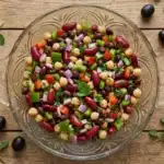 Three-Bean Salad: A Perfect Weeknight Treat 7 Three-bean salad in ornate glass serving bowl on rustic wooden table with chickpeas, kidney beans, black beans, diced bell peppers, red onions, and fresh herbs